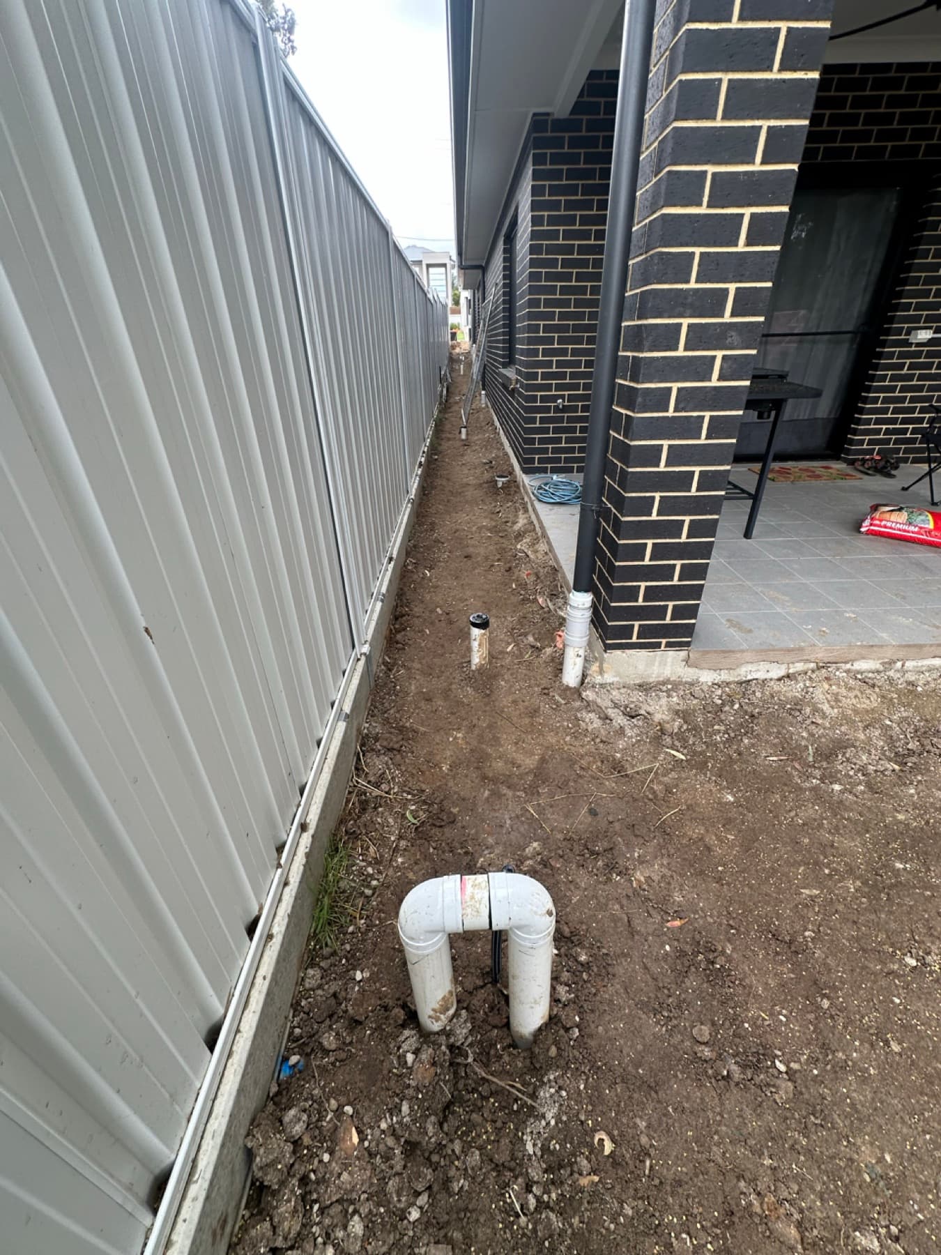 Narrow side passage between house and fence with two PVC pipe stubs installed along the ground