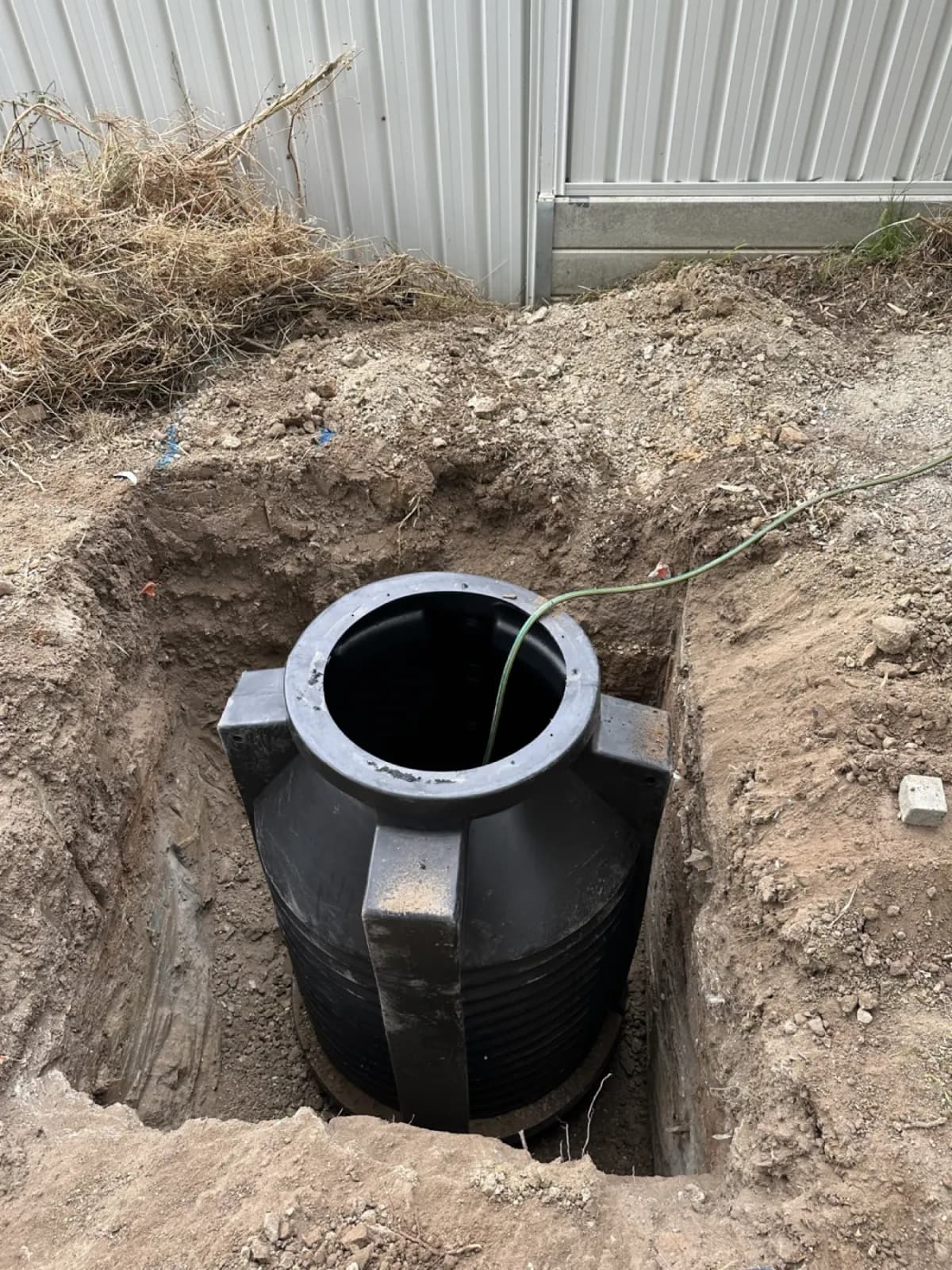 Black poly underground tank being lowered into an excavated pit beside a corrugated fence
