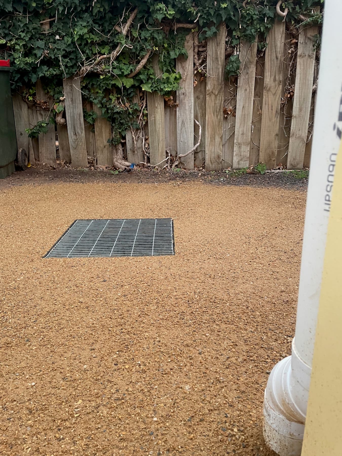 Square metal surface drainage grate installed flush in a gravel yard beside a downpipe